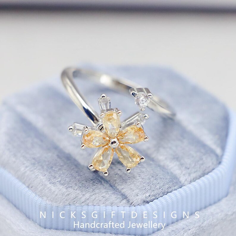 May include: A silver-toned ring featuring a flower design with yellow and clear gemstones. The flower has six petals and a central stone. The ring also has a small, clear gemstone on the band. The text "NICKS GIFTDESIGNS Handcrafted Jewellery" is visible.