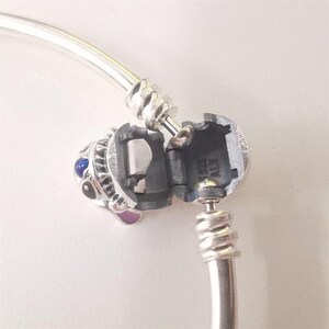 Puede incluir: Una pulsera plateada con un charm con un dise&ntilde;o detallado. El charm tiene detalles en azul y morado, y el cierre est&aacute; abierto, revelando el mecanismo interior. La pulsera est&aacute; sobre un fondo blanco.