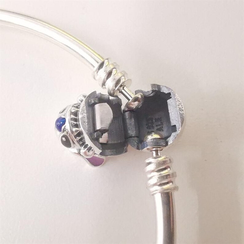 Puede incluir: Una pulsera plateada con un charm con un dise&ntilde;o detallado. El charm tiene detalles en azul y morado, y el cierre est&aacute; abierto, revelando el mecanismo interior. La pulsera est&aacute; sobre un fondo blanco.