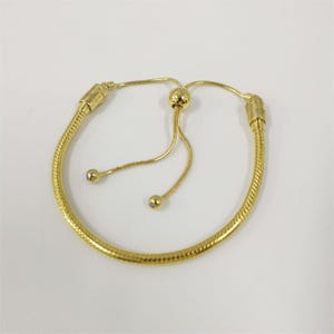 May include: A gold bracelet with a snake chain design. The bracelet has a sliding bead closure and two dangling gold beads. The bracelet is set against a white background.
