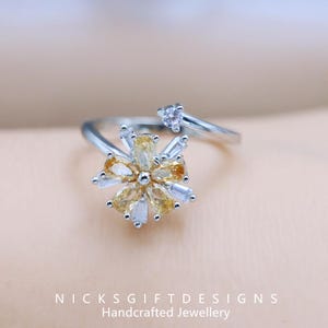 May include: A silver-toned ring featuring a flower design with yellow and clear gemstones. The flower has a central cluster of yellow stones surrounded by clear, baguette-cut stones. A small, clear gemstone accents the band.