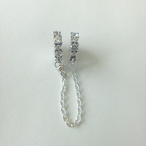 May include: A pair of silver-toned earrings with a chain. Each earring features three clear, square-cut stones. The earrings are connected by a delicate chain, creating a stylish and elegant accessory.