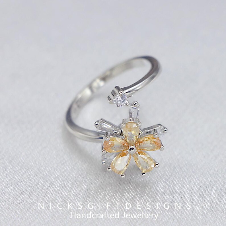 May include: A silver-toned ring featuring a flower design with yellow and clear gemstones. The flower has six yellow petal-shaped stones and is accented with clear, rectangular and round stones. The ring band is open and adjustable.