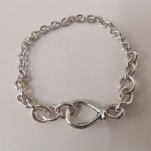 May include: A silver bracelet with a chain-link design. The bracelet features a large, oval-shaped clasp and is made of shiny, polished metal. The bracelet is designed to be worn around the wrist.
