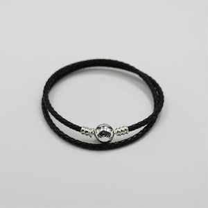 May include: A black braided leather bracelet with a silver clasp. The bracelet is made of two strands of black leather and has a round silver clasp in the centre. The bracelet is designed to be worn around the wrist.