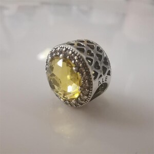 May include: A silver charm with a large, faceted yellow gemstone at the center. The charm has a detailed design with small, clear stones around the yellow gem and a patterned, openwork design on the sides. The word "ALE" is visible.