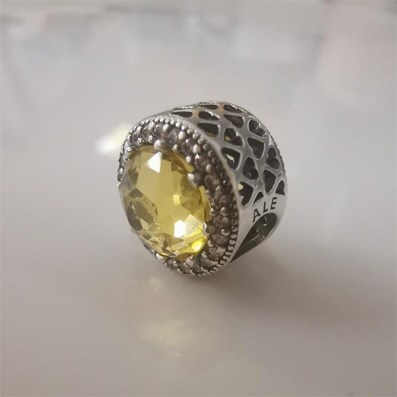 May include: A silver charm with a large, faceted yellow gemstone at the center. The charm has a detailed design with small, clear stones around the yellow gem and a patterned, openwork design on the sides. The word "ALE" is visible.