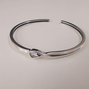 May include: A silver-toned cuff bracelet with a teardrop-shaped clear gemstone. The bracelet has a double-band design with a black accent on the inner band. The gemstone is set in a silver-toned setting.