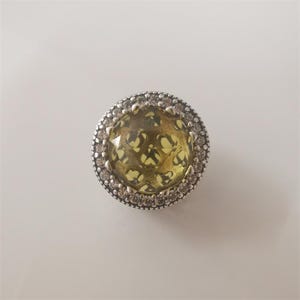May include: A round, silver-toned charm with a yellow-green center. The center has a cut-out design. The charm is surrounded by a ring of small, clear stones and a decorative silver border. Jewelry.