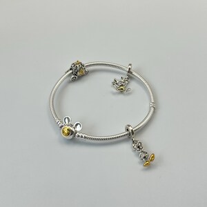 May include: Silver bracelet with multiple charms. The bracelet features a Mickey Mouse charm, a carriage charm, and a charm of a character with yellow accents. The bracelet is on a white background.