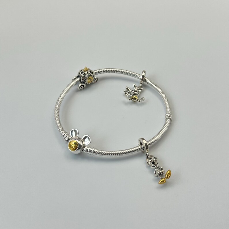 May include: Silver bracelet with multiple charms. The bracelet features a Mickey Mouse charm, a carriage charm, and a charm of a character with yellow accents. The bracelet is on a white background.
