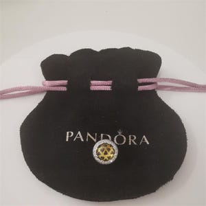 May include: A black velvet pouch with a pink drawstring and the word "PANDORA" in silver lettering. A silver charm with a yellow center and a heart pattern is in front of the pouch.