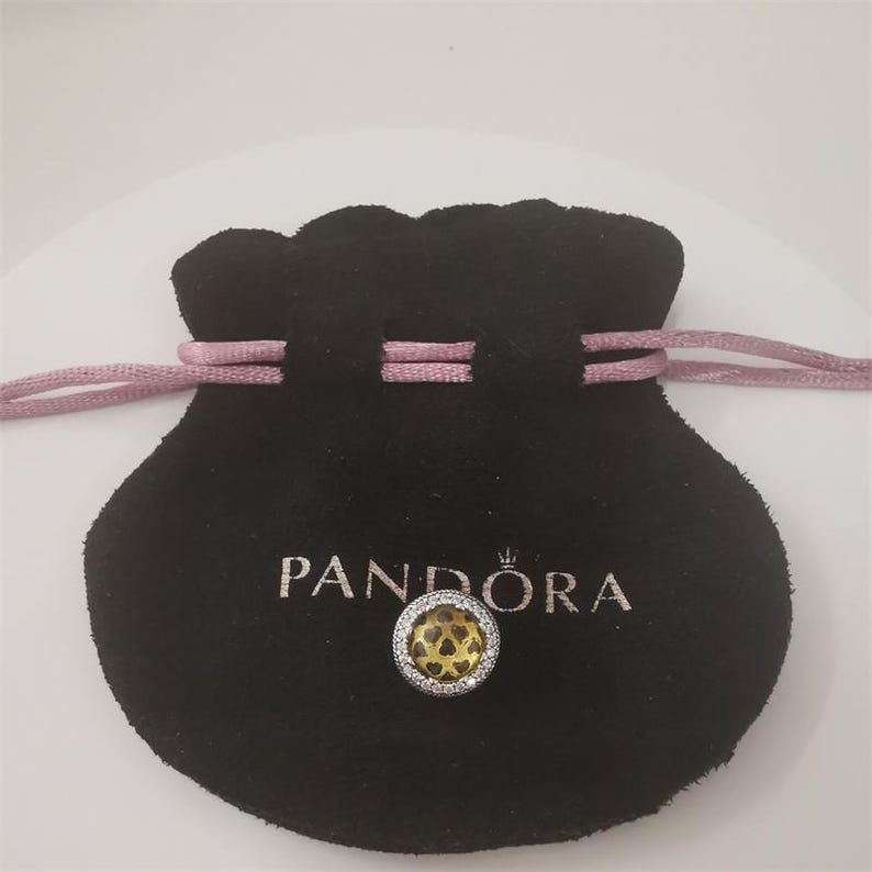 May include: A black velvet pouch with a pink drawstring and the word "PANDORA" in silver lettering. A silver charm with a yellow center and a heart pattern is in front of the pouch.