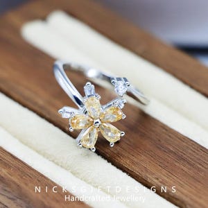 May include: A silver-toned ring featuring a flower design with yellow and clear gemstones. The flower is the focal point, with a heart-shaped accent on the band. The ring is displayed on a wooden surface. Text on the image reads: NICKSGIFTDESIGNS Handcrafted Jewellery.