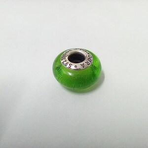 May include: A round, green glass bead with a silver-colored metal ring. The bead has a translucent appearance, and the metal ring is engraved with the word "PANDORA" and "S925". The bead is likely a charm for a bracelet or necklace.
