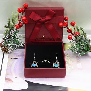 May include: A red gift box with a matching bow, open to reveal a pair of crab earrings and a ring. The earrings and ring are silver-toned with blue accents. Decorative sprigs with red berries and pine needles surround the box.