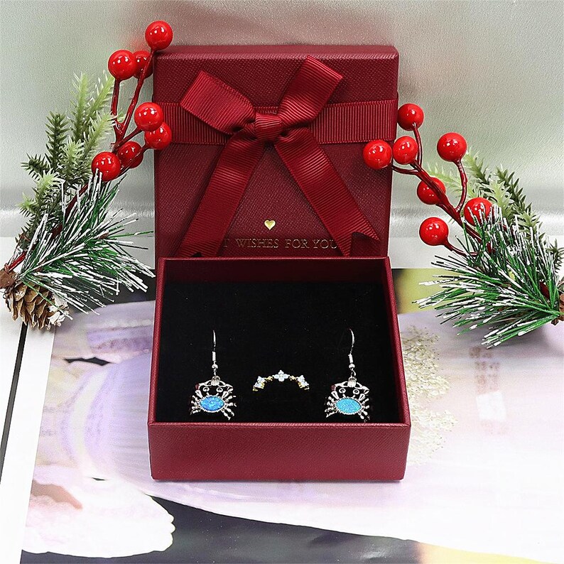 May include: A red gift box with a matching bow, open to reveal a pair of crab earrings and a ring. The earrings and ring are silver-toned with blue accents. Decorative sprigs with red berries and pine needles surround the box.