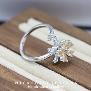 May include: A silver ring with a flower design. The flower has yellow and clear gemstones. The ring is on a wooden surface. The text "NICKSGIFTDESIGNS Handcrafted Jewellery" is at the bottom of the image.
