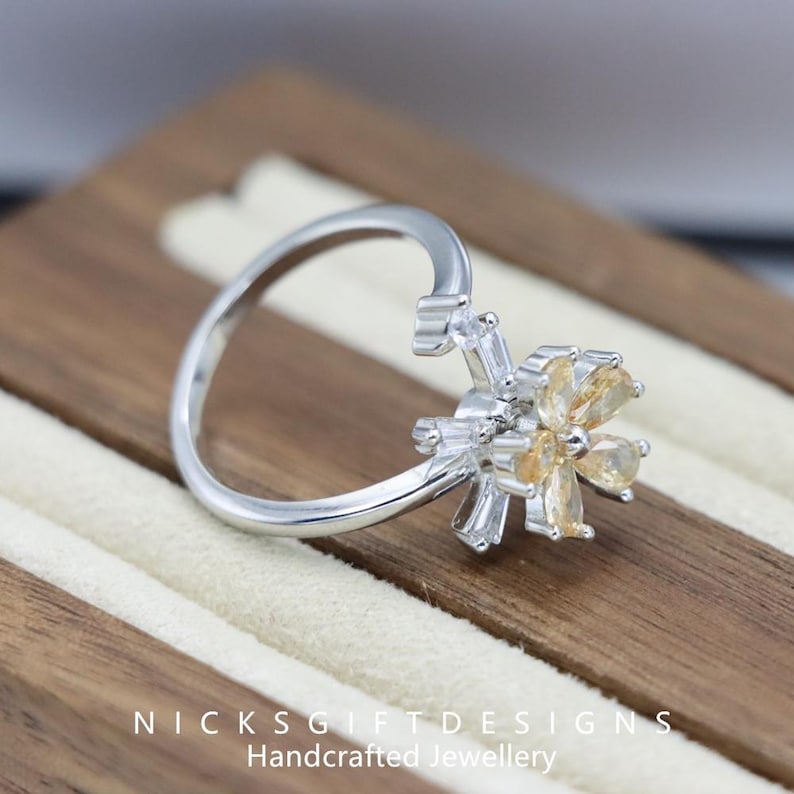 May include: A silver ring with a flower design. The flower has yellow and clear gemstones. The ring is on a wooden surface. The text "NICKSGIFTDESIGNS Handcrafted Jewellery" is at the bottom of the image.