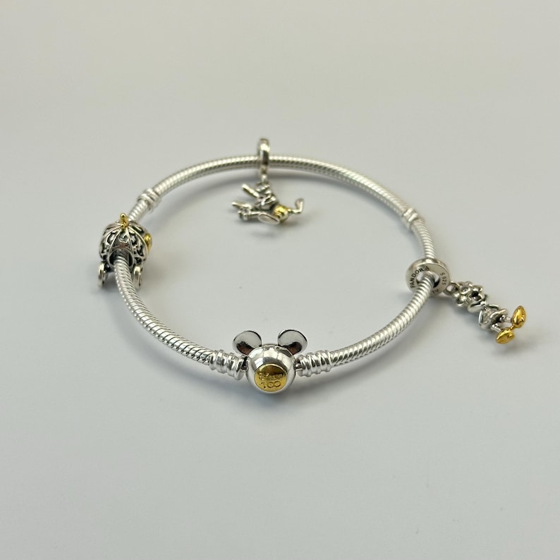 May include: A silver bracelet with multiple charms. Charms include a crown, a Mickey Mouse head, and a Mickey Mouse figure with gold accents. The bracelet is on a white surface.