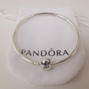 Puede incluir: Una pulsera Pandora plateada con un charm de un personaje con ojos azules. La pulsera est&aacute; sobre una bolsa blanca con el logo de Pandora y el texto "Love at first bite".