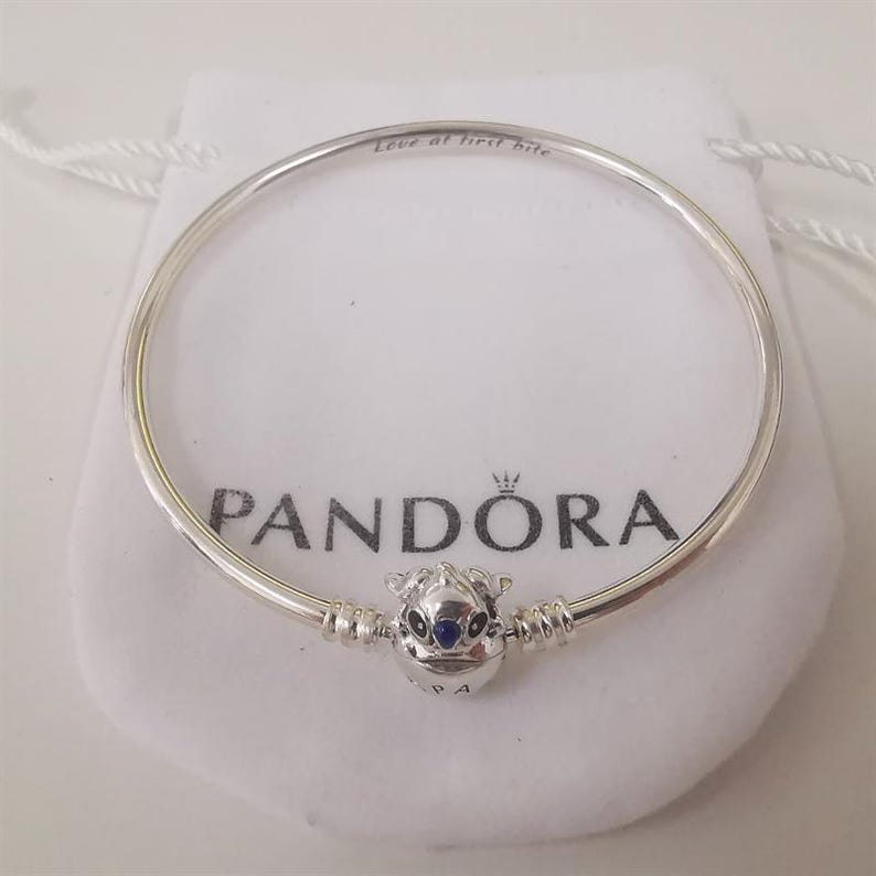 Puede incluir: Una pulsera Pandora plateada con un charm de un personaje con ojos azules. La pulsera est&aacute; sobre una bolsa blanca con el logo de Pandora y el texto "Love at first bite".