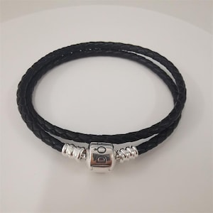 May include: A black leather wrap bracelet with a braided design. The bracelet has a silver-coloured clasp with a brand logo. The bracelet is designed to wrap around the wrist twice.