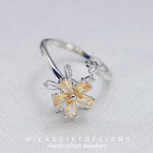 May include: A silver-toned ring featuring a flower design with yellow and clear gemstones. The flower has a central cluster of yellow pear-shaped stones surrounded by clear, rectangular and round stones. The ring band is open and adjustable.