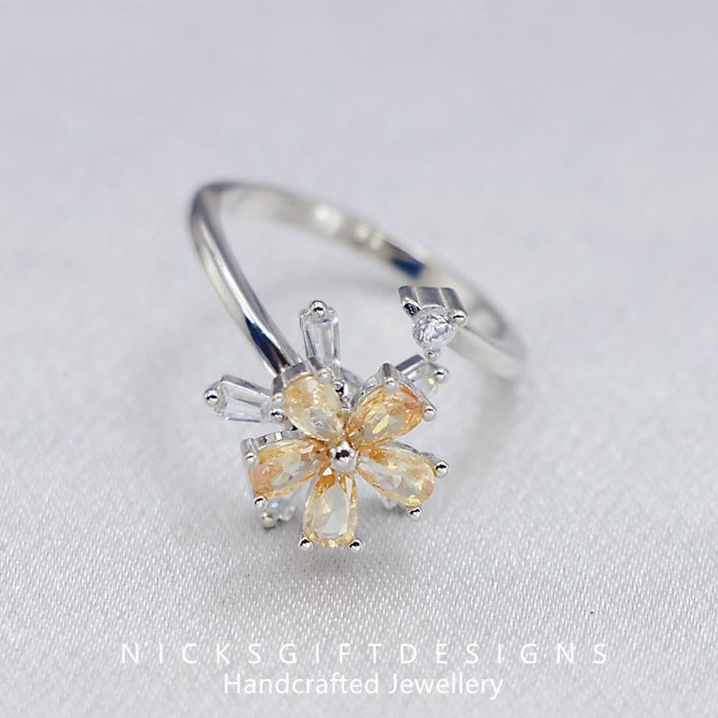 May include: A silver-toned ring featuring a flower design with yellow and clear gemstones. The flower has a central cluster of yellow pear-shaped stones surrounded by clear, rectangular and round stones. The ring band is open and adjustable.