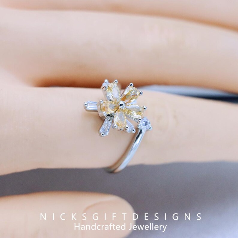 May include: A silver ring featuring a flower design with yellow and clear gemstones. The ring's band curves gracefully, with a small clear gemstone accent. The text "NICKS GIFT DESIGNS Handcrafted Jewellery" is visible at the bottom.