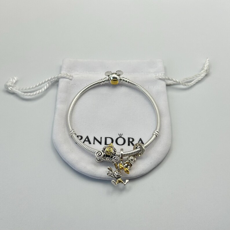 May include: A silver Pandora bracelet with a Mickey Mouse clasp and charm. The bracelet features a gold-colored Mickey Mouse head clasp and several charms, including a gold-colored carriage and a charm with a cartoon character.