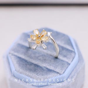 May include: A silver ring with a flower design, featuring yellow and clear gemstones. The flower has six petals and is set on a delicate band. The ring is displayed in a blue velvet ring box. The text "NICKSGIFTDESIGNS Handcrafted Jewellery" is visible.