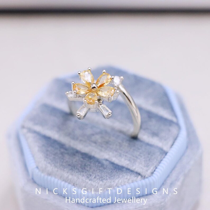 May include: A silver ring with a flower design, featuring yellow and clear gemstones. The flower has six petals and is set on a delicate band. The ring is displayed in a blue velvet ring box. The text "NICKSGIFTDESIGNS Handcrafted Jewellery" is visible.