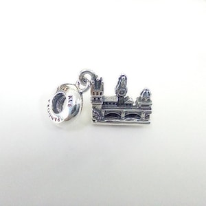 May include: A silver charm featuring a detailed miniature of a bridge and buildings. The charm has a loop for attaching to a bracelet or necklace. The charm is made of silver and has the word "Pandora" and "ALE" engraved on it.