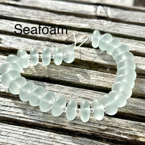 May include: A seafoam green bracelet composed of oval beads. The beads are linked with a thin, transparent string. The word "Seafoam" is written in black text at the top of the image. The bracelet is displayed on a weathered wooden surface.
