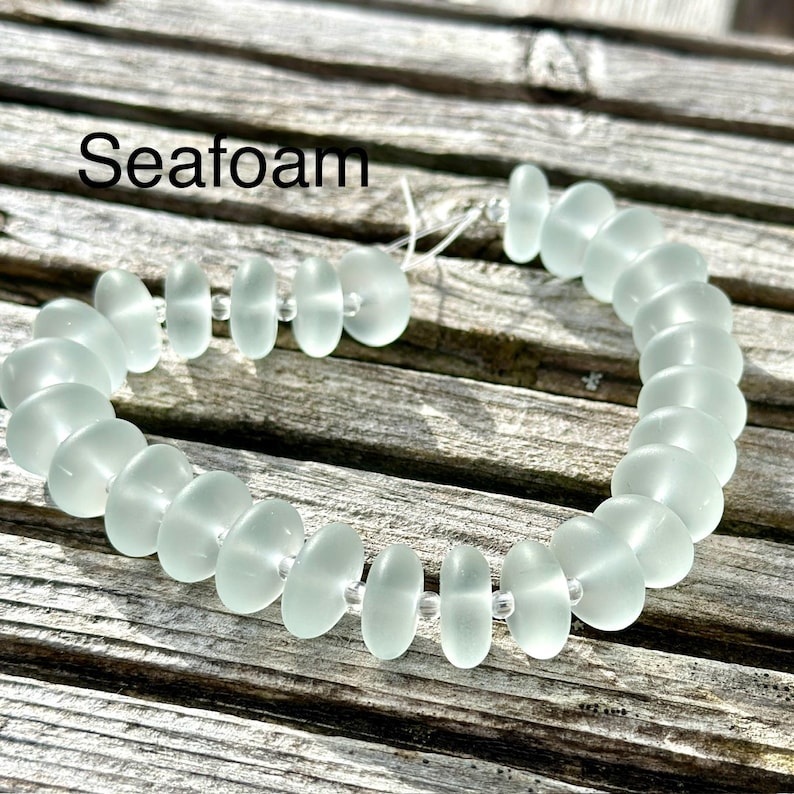 May include: A seafoam green bracelet composed of oval beads. The beads are linked with a thin, transparent string. The word "Seafoam" is written in black text at the top of the image. The bracelet is displayed on a weathered wooden surface.