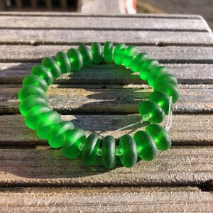May include: A bright green bracelet made from rounded sea glass beads. The beads are strung on a clear cord, forming a circle. The bracelet is shown on a weathered wooden surface, with natural light highlighting the colour. The beads are approximately 1 cm in diameter.