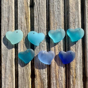 Peut inclure: Sept morceaux de verre de mer en forme de cœur, dans différentes nuances de bleu et de turquoise. Chaque pièce a un petit trou en haut. Le verre de mer est disposé sur une surface en bois vieilli.
