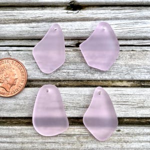 May include: Four pale purple sea glass pendants with a frosted finish. Each pendant has a unique shape and a small hole at the top for stringing. The pendants are displayed on a weathered wooden surface.