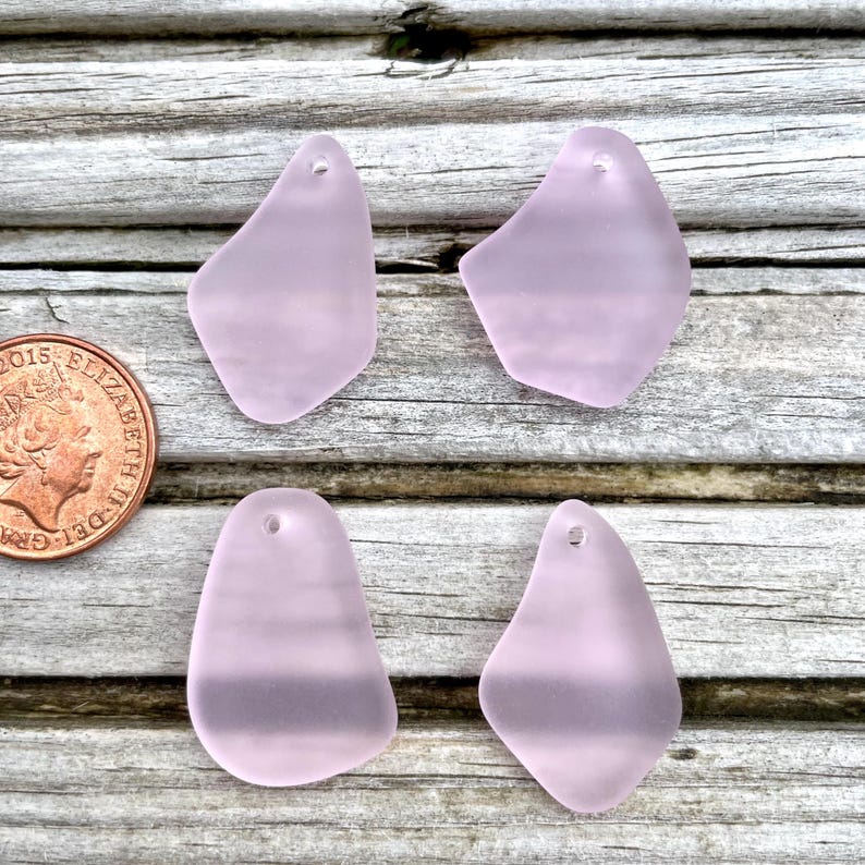 May include: Four pale purple sea glass pendants with a frosted finish. Each pendant has a unique shape and a small hole at the top for stringing. The pendants are displayed on a weathered wooden surface.