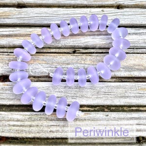 May include: A periwinkle-coloured beaded bracelet. The bracelet features flat, oval-shaped beads strung together with clear beads. The word "Periwinkle" is written in a white font at the bottom of the image. The beads have a soft, matte finish.