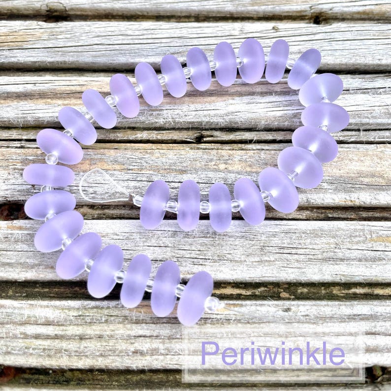 May include: A periwinkle-coloured beaded bracelet. The bracelet features flat, oval-shaped beads strung together with clear beads. The word "Periwinkle" is written in a white font at the bottom of the image. The beads have a soft, matte finish.