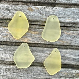 May include: Four pale yellow sea glass pendants with drilled holes. The pendants have irregular shapes and a frosted appearance, ideal for jewellery making. Each pendant is a unique shape.