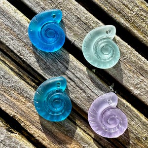 May include: Four sea glass spiral pendants in blue, teal, and purple hues. Each pendant has a small hole for threading. They are displayed on a weathered wooden surface, highlighting their translucent nature and distinctive forms.