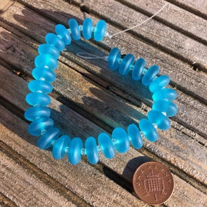 May include: A string of light blue, oval glass beads, connected with clear spacers, forming a circular shape. A penny is in the lower right corner for size reference. The beads are approximately 1 cm in diameter.