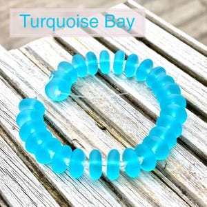 May include: A bracelet composed of translucent, light blue, oval beads threaded on a clear cord. The beads are arranged in a circular design. The text "Turquoise Bay" is displayed in a light blue font in a white rectangle.