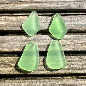 May include: Four sea glass pendants in a soft, translucent green hue. Each piece has a smooth, rounded shape with a small hole at the top for stringing. The pendants are displayed on a weathered wooden surface.