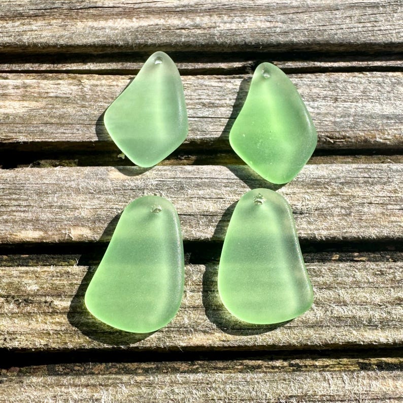 May include: Four sea glass pendants in a soft, translucent green hue. Each piece has a smooth, rounded shape with a small hole at the top for stringing. The pendants are displayed on a weathered wooden surface.
