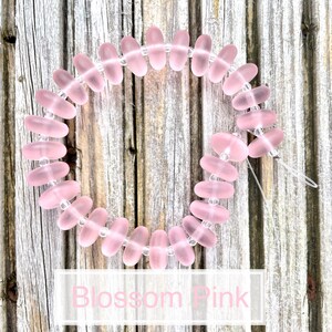 May include: A string of translucent pink oval beads with small clear spacer beads. The beads are strung together on a clear string. The text "Blossom Pink" is displayed in a white rectangle.