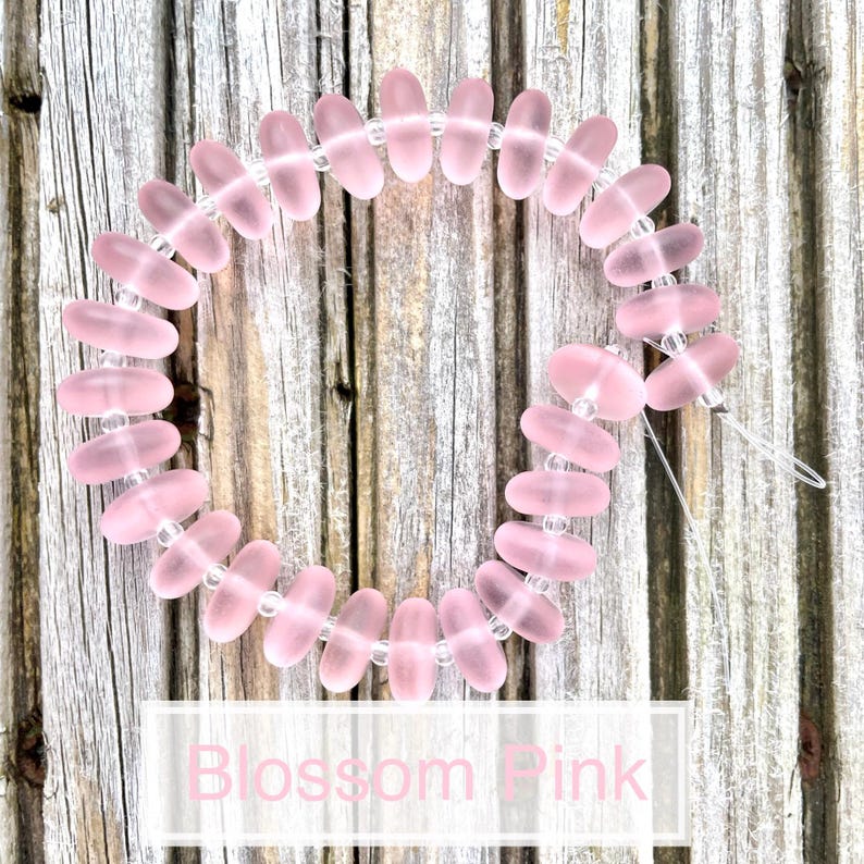 May include: A string of translucent pink oval beads with small clear spacer beads. The beads are strung together on a clear string. The text "Blossom Pink" is displayed in a white rectangle.